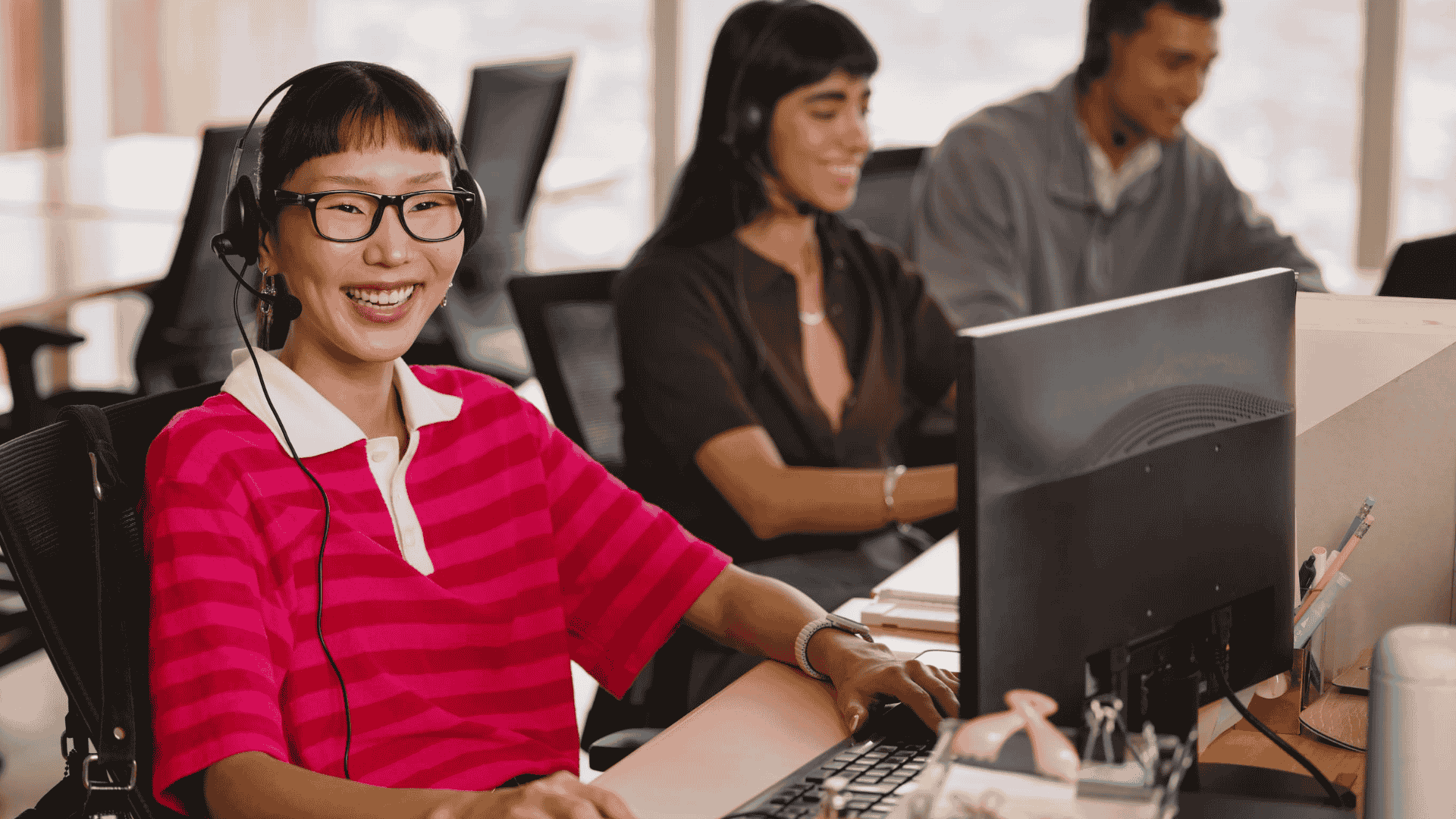 A smiling agent in a pink striped shirt and headset sits at a computer.