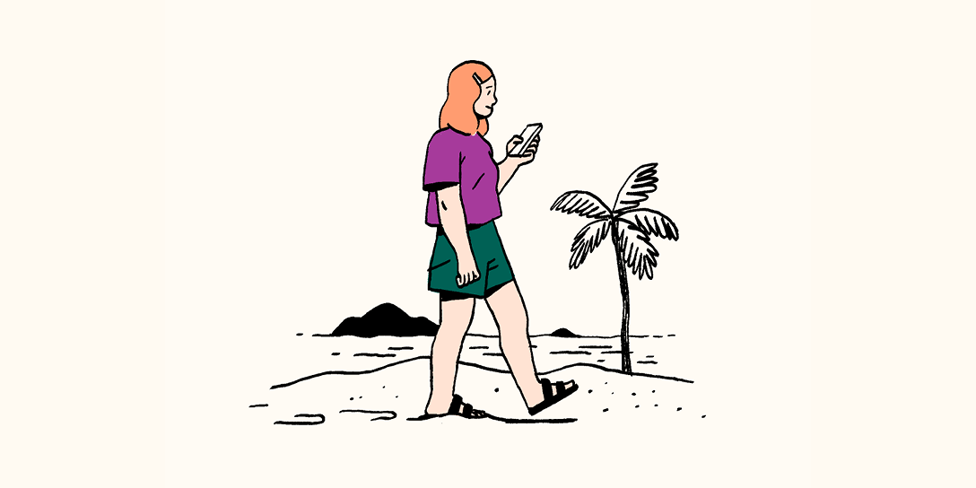 A woman with red hair walks on the beach while using her phone.